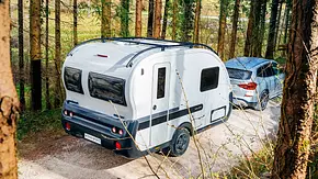 Der Adria Action Sports 391 LH (2025) von schräg hinten rechts von einem Auto gezogen auf einer Schotterstraße zwischen Bäumen. - Foto: Adria
