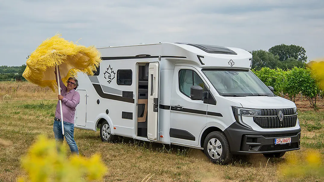 Mann mit gelbem Sonnenschirm steht neben einem weißen Ahorn Camp T 690 First Edition Wohnmobil auf einer Wiese. - Foto: Hardy Mutschler