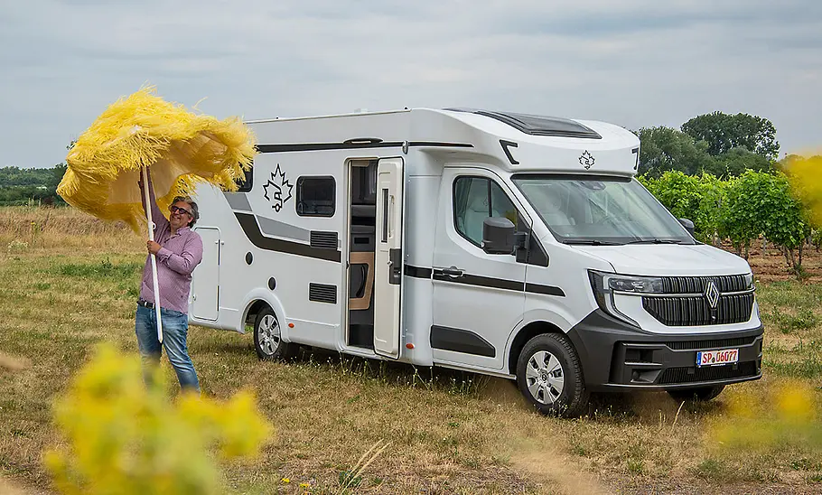 Ahorn Camp T 690 First Edition (2025) Mann mit gelbem Sonnenschirm steht neben einem weißen Ahorn Camp T 690 First Edition Wohnmobil auf einer Wiese.