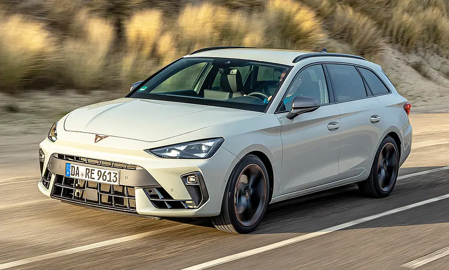 Cupra Leon Sportstourer Der Cupra Leon Sportstourer fährt auf einer Straße an einer Düne entlang, von vorne links fotografiert.