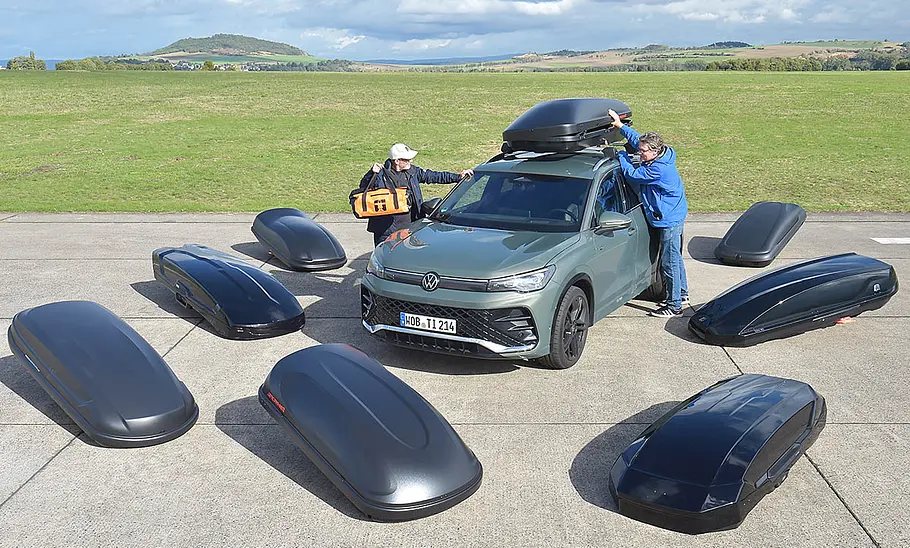 Dachboxen-Test Überblicksbild mit sieben Dachboxen auf der Teststrecke liegend, mit dem Testfahrzeug inklusive montierter Dachbox und zwei Personen in der Mitte des Bildes.