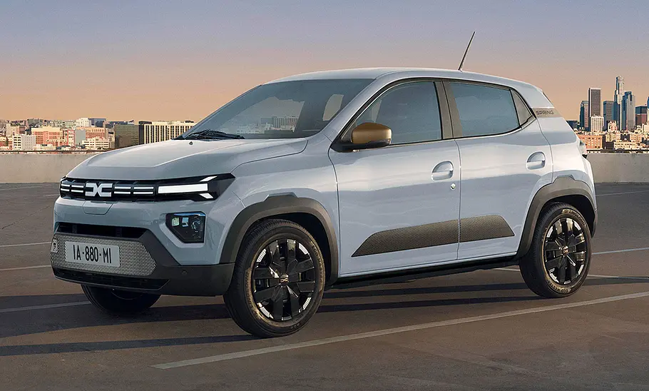 Dacia Spring Facelift (2024) Das Dacia Spring Facelift (2024) steht vor urbanem Hintergrund auf einem Parkplatz, von vorne links fotografiert.