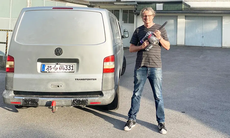 Die Außenaufnahme zeigt einen Tester mit dem Dyson Akku-Staubsauger Car & Boat V8 in der Hand vor einem silbernen VW Bus.