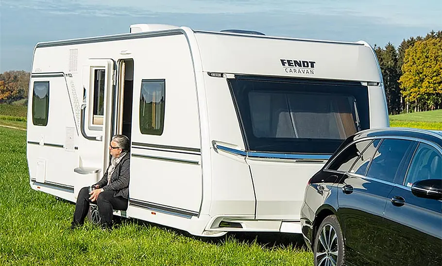 Fendt Caravan Apero 560 SKM (2024) Fendt Caravan Apero 560 SKM (2024) hängt an einem Auto und parkt auf einer Wiese. In der Eingangstür zum Caravan sitzt eine Person.