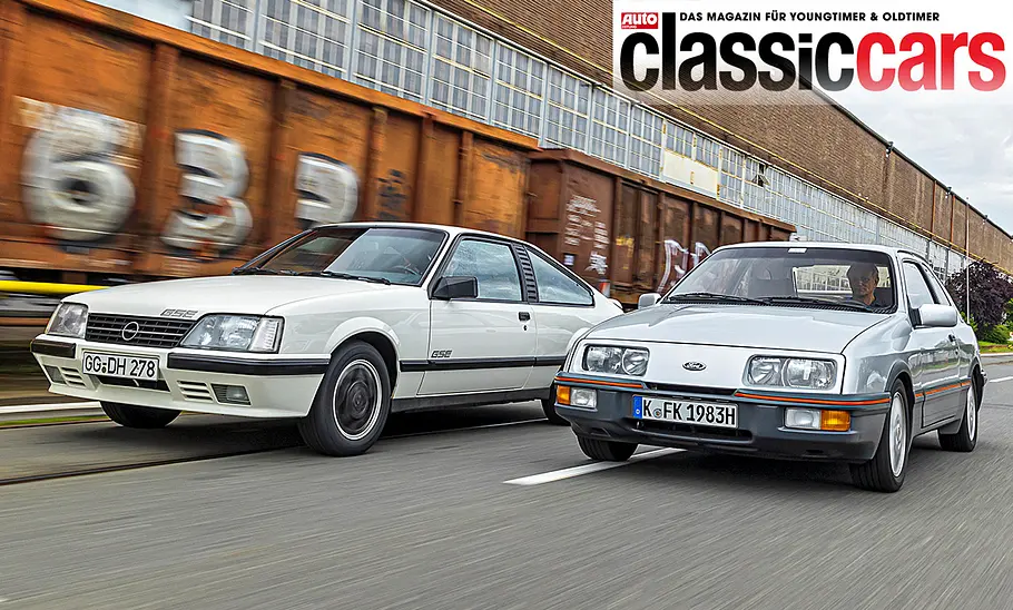 Opel Monza GSE/Ford Sierra XR4i Opel Monza GSE und Ford Sierra XR4i fahren nebeneinander durch ein Industriegebiet, von vorne fotografiert.