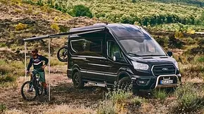 Die Außenaufnahme zeigt den Karmann Dexter 560 4x4 (2025) statisch von schräg vorne links. Vor dem Wohnmobil steht eine Person auf einem Mountainbike, die Markise ist ausgefahren. - Foto: Karmann Mobil