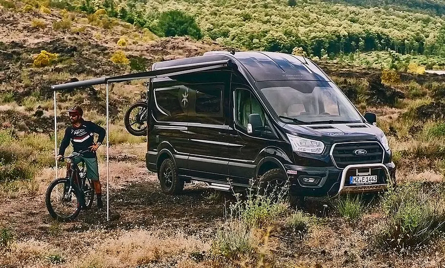Die Außenaufnahme zeigt den Karmann Dexter 560 4x4 (2025) statisch von schräg vorne links. Vor dem Wohnmobil steht eine Person auf einem Mountainbike, die Markise ist ausgefahren.