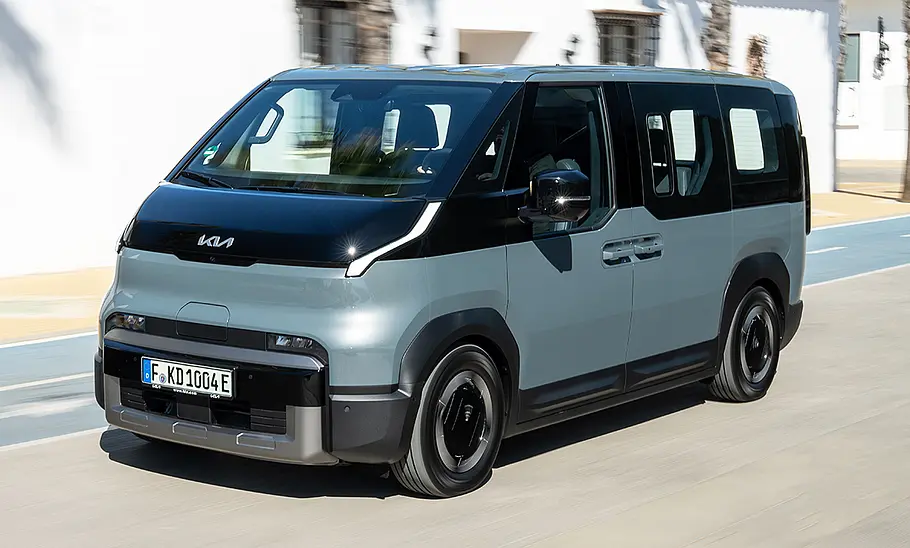 Der Kia PV5 Passenger (2025) fährt durch eine urbane Umgebung, von vorne links fotografiert.
