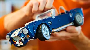 Die Lego Icons Shelby Cobra von schräg vorne links. Eine Person (im Bild nur unscharf im Hintergrund zu sehen) hält das Modellauto nah an die Kamera. - Foto: Lego