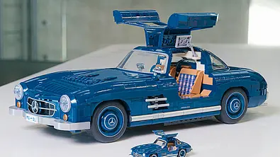 Das Bild zeigt den Mattel Brick Shop Mercedes 300 SL von schräg vorne links mit geöffneten Flügeltüren. Vor dem Klemmbaustein-Auto steht eine Diecast-Miniatur in kleinerem Maßstab. - Foto: Mattel