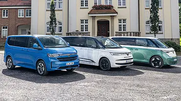 VW Caravelle, Multivan und ID. Buzz stehen nebeneinander auf einem Schotterparkplatz. - Foto: VW