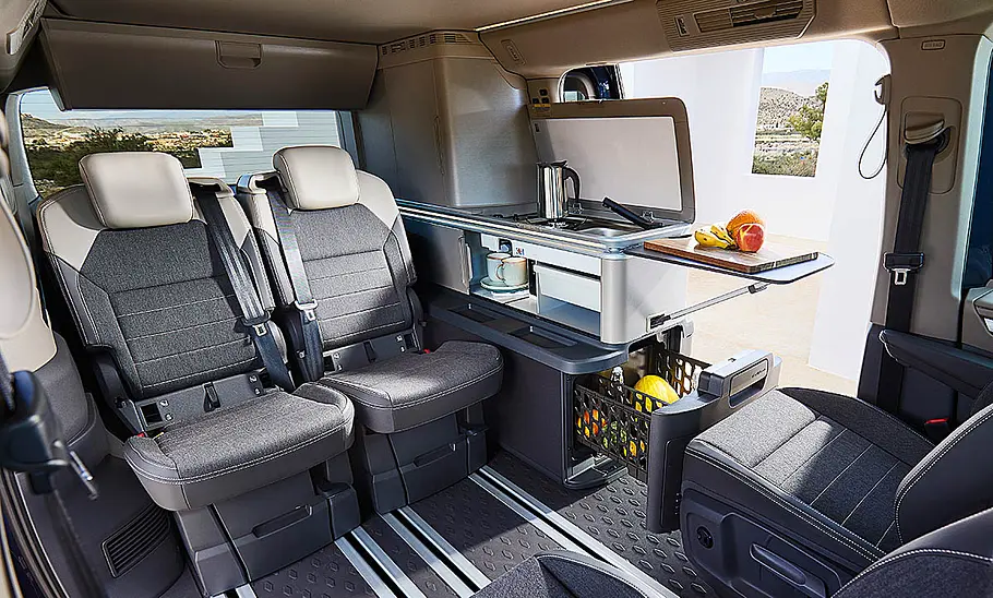 Das Bild zeigt den Wohnraum des VW California Ocean (2024). Zu sehen ist der Wohnraum mit voller Bestuhlung und Seitenküche mit geöffnetem Kühlschrank.