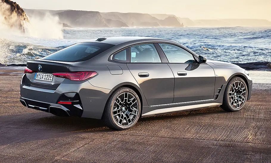 Das BMW i4 Facelift (2024) steht vor einem Küsten- und Gebirgspanorama, von hinten rechts fotografiert.