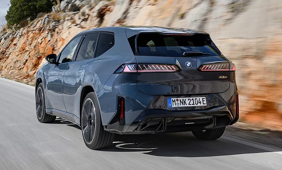 Der BMW iX3 fährt auf einer Bergstraße, von hinten links fotografiert.