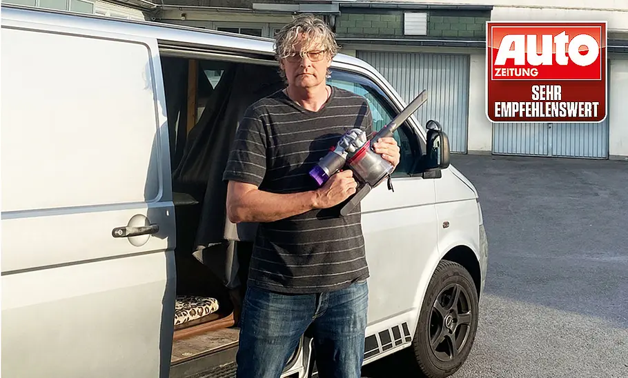 Die Außenaufnahme zeigt einen Tester mit dem Dyson Akku-Staubsauger Car & Boat V8 in der Hand vor der geöffneten Tür eines silbernen VW Bus.