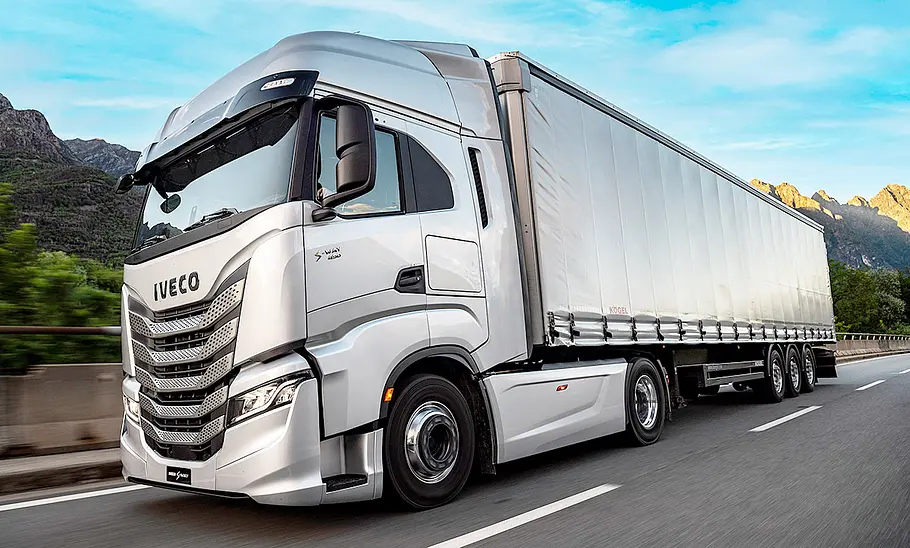 Der Iveco S-Way (2024) fährt über eine Landstraße mit malerischem Bergpanorama, von vorne links fotografiert.