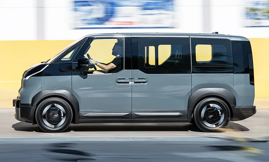 Der Kia PV5 Passenger (2025) fährt durch eine urbane Umgebung, von links fotografiert.