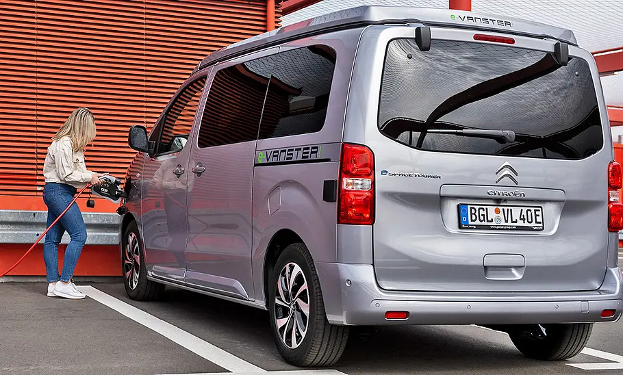 Pössl Vanster Das Bild zeigt den Pössl Vanster in der Elektrovariante e-Vanster statisch von schräg hinten links. Eine Person schließt das Auto zum Laden per Ladekabel an der Buchse im vorderen linken Kotflügel an.