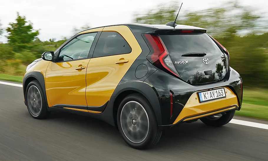 Toyota Aygo X Facelift (2025) fährt auf einer Landstraße, Ansicht von hinten links.