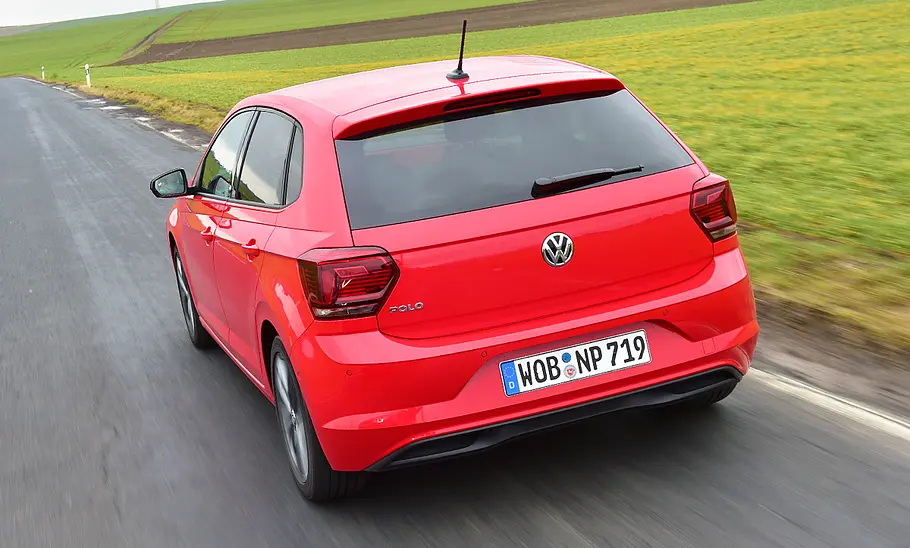 Der VW Polo (2017) fährt über eine Straße, im Hintergrund ist eine Wiese zu sehen. Von hinten links fotografiert.