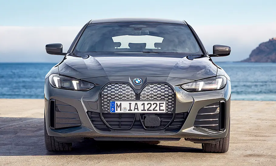 Das BMW i4 Facelift (2024) steht vor einem Küsten- und Gebirgspanorama, von vorne fotografiert.