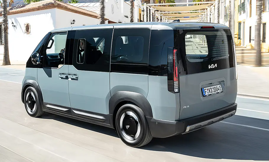 Der Kia PV5 Passenger (2025) fährt durch eine urbane Umgebung, von hinten links fotografiert.
