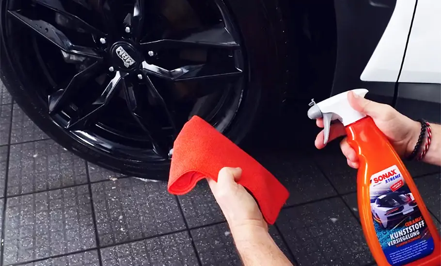 Rad und Radlauf eines Autos mit Händen, die die Sonax Kunststoffversiegelung und ein Microfasertuch halten.