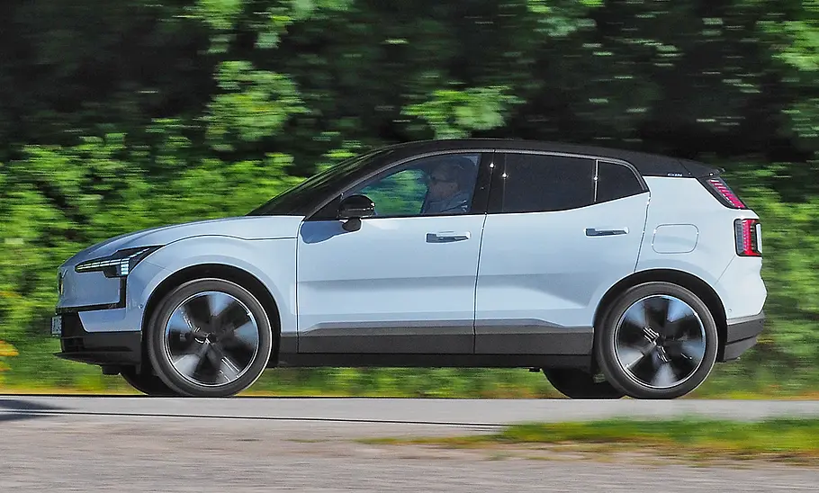 Der Volvo EX30 Single Motor Extended Range Ultra fährt von rechts nach links, der Hintergrund ist unscharf und zeigt grüne Gebüsche und Bäume.