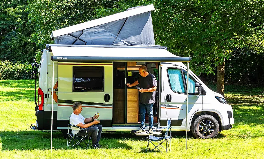 Der Weinsberg CaraBus 540 MQ (2025) statisch von der rechten Seite. Die Markise ist ausgefahren, das Aufstelldach geöffnet, eine Person sitzt auf einem Campingstuhl unter der Markise, eine weitere Person steht in der geöffneten Schiebetüre.