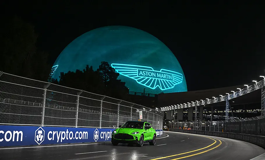 Der Aston Martin DBX auf einer leeren Straße fahrend, im Hintergrund sieht man groß das Markenlogo.