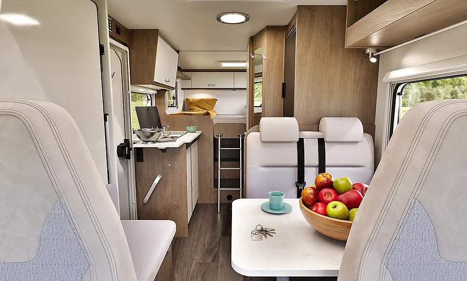 Wohnraum des Carado V347 vom Fahrerhaus aus gesehen, Tisch mit Obstschale und Tasse, dahinter Sitzbank, Küche mit Kochfeld und Spüle sowie Zugang zu den Betten im Heck.