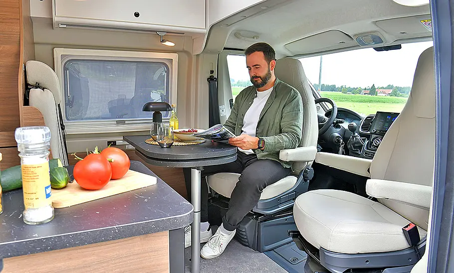 Das Bild zeigt die Sitzgruppe des Corigon CV 60 D (2025) durch die geöffnete Schiebetüre mit Blick in Richtung Cockpit fotografiert. Eine Person sitzt auf dem in Richtung Wohnraum gedrehten Fahrersitz.