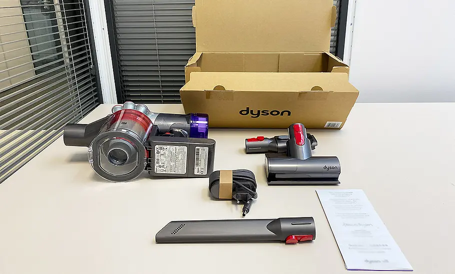 Die Innenaufnahme zeigt die Einzelteile des Lieferumfangs des Dyson Akku-Staubsaugers Car & Boat V8 und dem Lieferkarton auf einem Tisch liegen.
