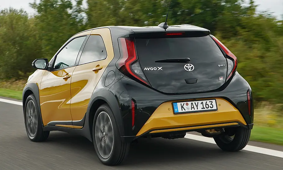 Toyota Aygo X Facelift (2025) fährt auf einer Landstraße, Ansicht von hinten links.