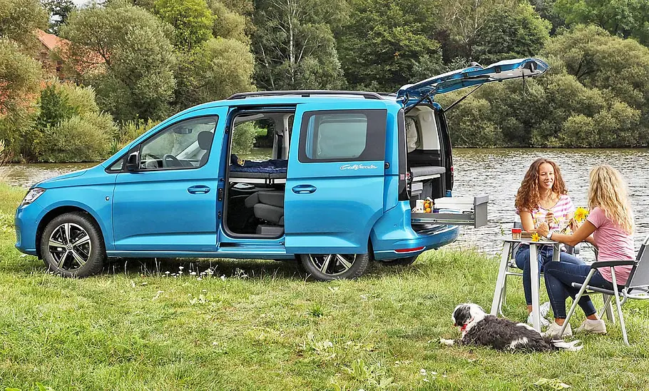 VW Caddy California (2020) auf einer Wiese am See geparkt und mit offenstehenden Türen, zwei Personen an einem Campingtisch im Vordergrund