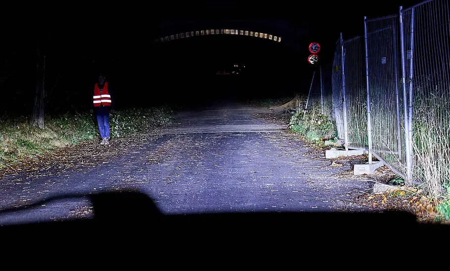 Nächtliche Landstraße mit Brücke und Fußgänger, die von Autoscheinwerfern angeleuchtet werden.