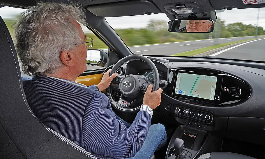 Toyota Aygo X Facelift (2025) fährt auf einer Landstraße. Man blickt von der Rückbank aus auf das Cockpit, den Fahrer und durch die Fontscheibe auf die Straße.