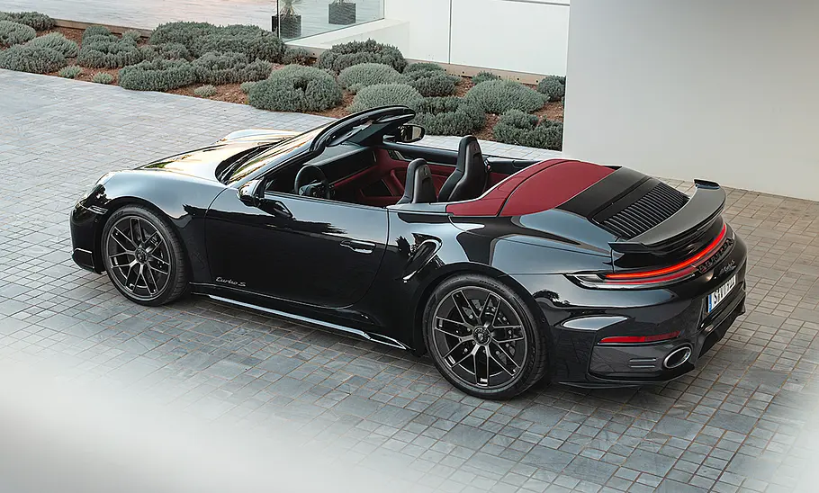 Das Porsche 911 Turbo S Cabrio Facelift (2025) steht mit offenem Dach vor einem Haus, von hinten links fotografiert.