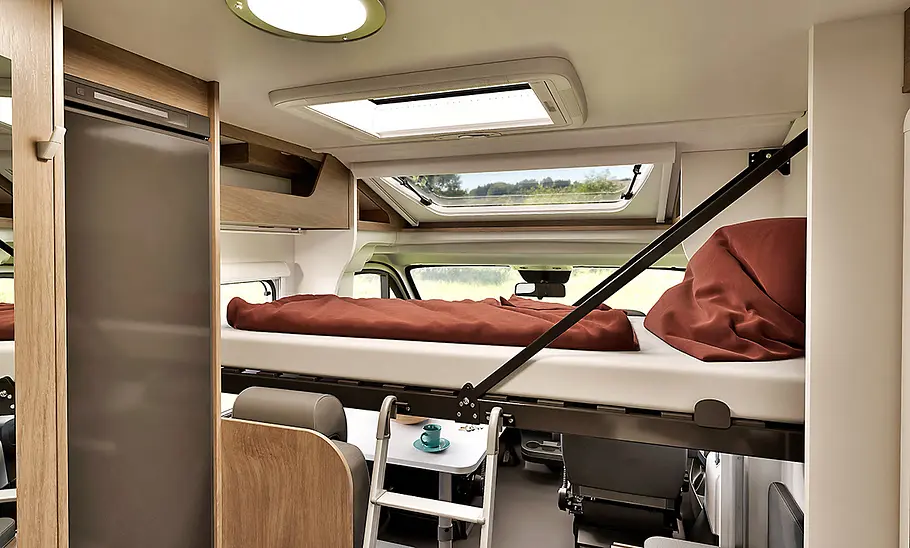 Carado V347 (2025) Innenansicht des Carado V347 mit abgesenktem Clever-Lift-Bett über der Sitzgruppe, braune Bettwäsche, Leiter für den Einstieg, großes Dachfenster.