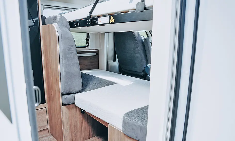 Carado V347 (2025) Innenraum mit abgesenktem Clever-Lift-Bett, darunter umgebaute Sitzgruppe als weiterer Schlafplatz, Polster in grauer Farbe, Blick in Richtung Fahrerhaus.
