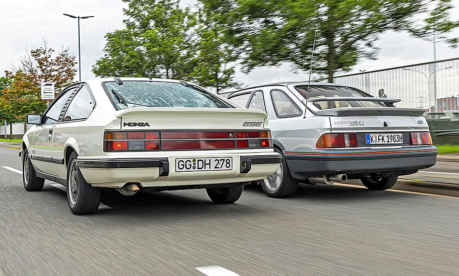 Opel Monza GSE/Ford Sierra XR4i Opel Monza GSE und Ford Sierra XR4i fahren nebeneinander durch ein Industriegebiet, von hinten fotografiert.