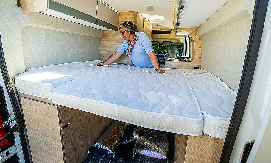 Die Innenaufnahme zeigt das Heck-Doppelbett des Forster Livin' Up Pro FV 599 (2025) durch die geöffneten Hecktüren. Eine Person misst die Länge des Betts.