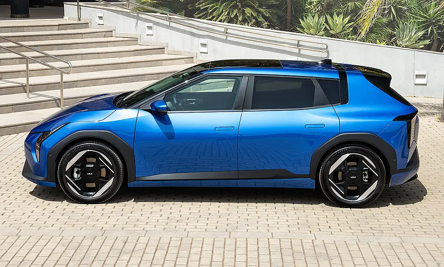 Kia EV4 (2025) Der Kia EV4 (2025) steht auf einem gepflasterten Platz vor einer Außentreppe, von links fotografiert.