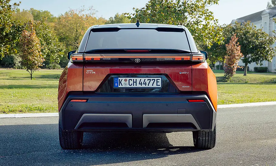 Toyota C-HR+ (2025) Der Toyota C-HR+ (2025) steht auf einer Straße im Grünen, von hinten fotografiert.