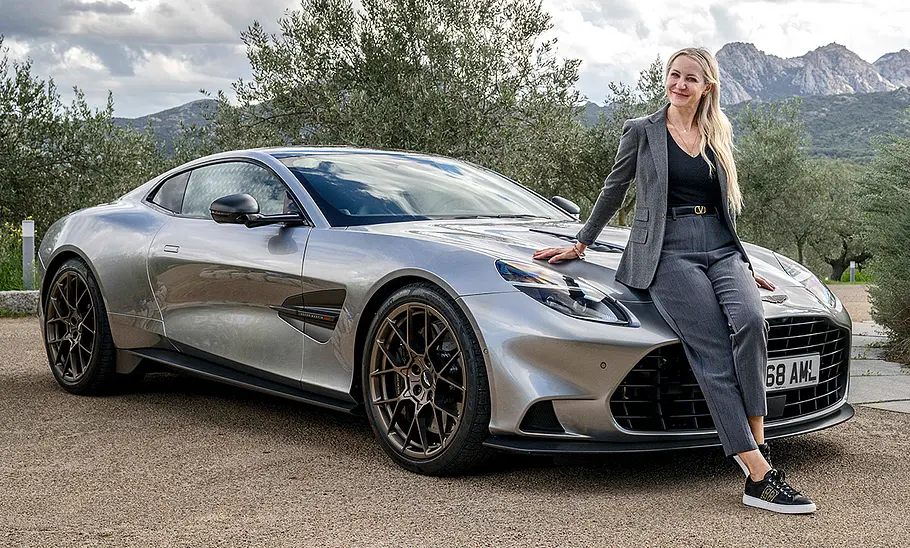 Stehender Aston Martin Vanquish (2024) mit Blick schräg von der linken Seite. AUTO ZEITUNG-Redakteurin Adele Moser steht an die Front angeleht vor dem Fahrzeug.