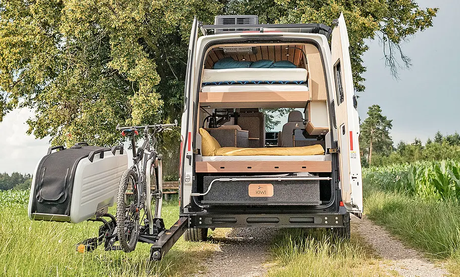 Kiwi Aotearoa (2024) Kiwi Aotearoa (2024) steht mit weggeklapptem Fahrradträger und Koffer am Heck auf einem Feldweg. Ansicht von hinten, die Hecktüren sind geöffnet.