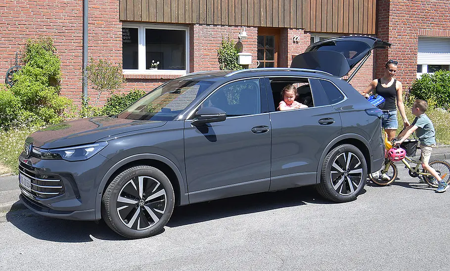 VW Tiguan Der VW Tiguan steht vor einem Wohnhaus, Ansicht des Wagens von vorne links. Ein Kind schaut aus dem rechten hinteren Fenster, an der geöffneten Kofferraumklappe steht die Mutter und ein Kind mit Fahrrad.
