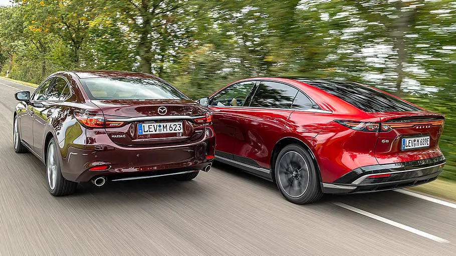 Mazda6 und Mazda6e fahren nebeneinander auf einer Landstraße, von hinten links fotografiert.