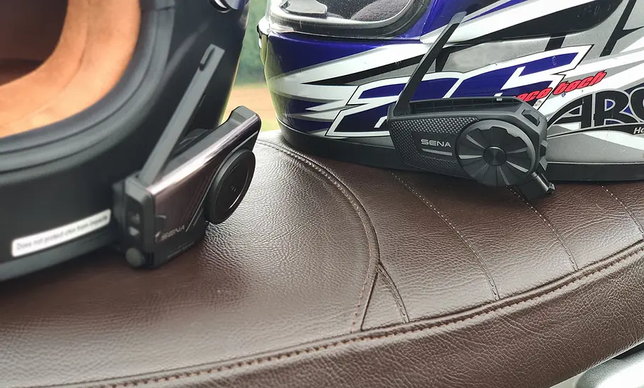 Sena 60S und Spider ST1 an jeweils einem Helm montiert, beide Helme liegen auf der Sitzbank eines Motorrads. Nahaufnahme der Intercom-Systeme.