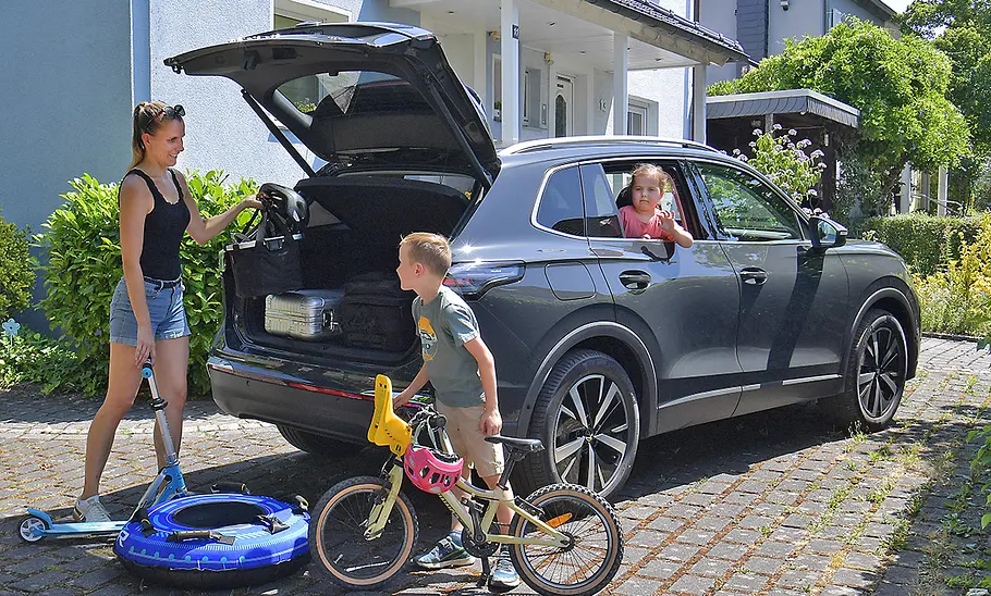 VW Tiguan Der VW Tiguan steht vor einem Wohnhaus, Ansicht des Wagens von hinten rechts. Ein Kind schaut aus dem rechten hinteren Fenster, an der geöffneten Kofferraumklappe steht die Mutter und recht einem Kind mit Fahrrad einen Fahrradhelm.